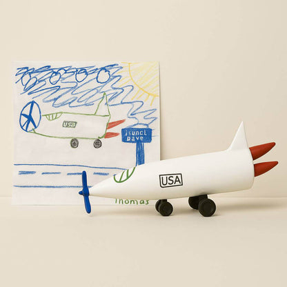 Imaginative crayon drawing of a USA-marked airplane, transformed into a detailed OstanesKids figurine with red rocket boosters and a blue propeller. A delightful handmade gift for creative kids.